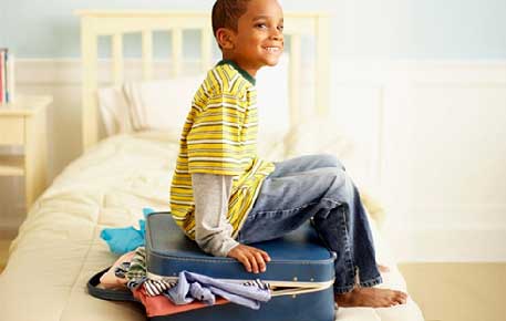A child smiling and sitting on a suitcase on a bed. The suitcase has clothing spilling out of it.