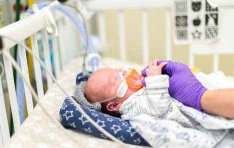 A baby in the intensive care unit holding the gloved hand of an adult healthcare provider.