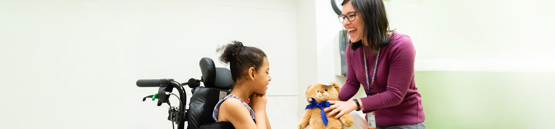 A healthcare provider hands a teddy bear to a young girl in a wheelchair