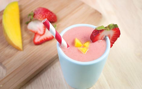 A pink strawberry smoothie in a white cup with a red and white striped straw. There is a cut piece of strawberry on the edge of the cup and yellow fruit chunks in the smoothie.